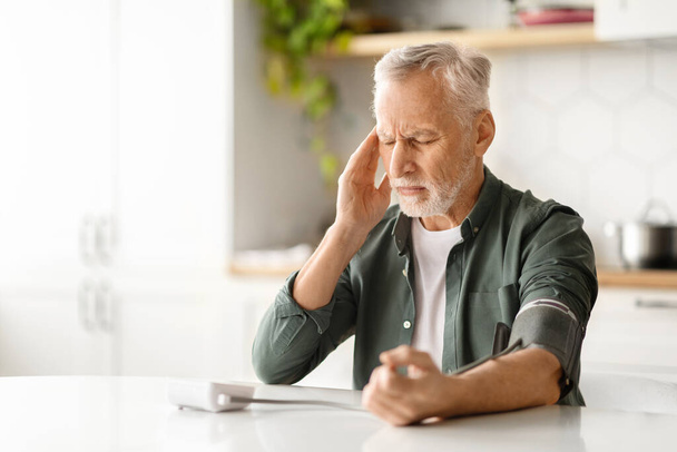 Aggódó idős férfi használja a felkar vérnyomás monitor, miközben ül a konyhaasztalnál, idős úriember rosszul érzi magát otthon, nézi eszköz, egészségügyi problémák - Fotó, kép