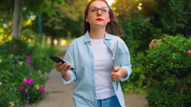 Boldog nő laptoppal és okostelefonnal a kezében sétál a virágzó kertben vagy a zöld utcában munka után nap. Lassú mozgás. - Felvétel, videó