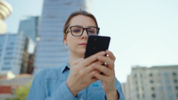 Gözlüklü beyaz kadın şehrin caddesinde dikiliyor ve akıllı telefon kullanıyor. Arka planda gökdelenler var. İletişim, iş günü, yoğun yaşam konsepti. Yavaş çekim - Video, Çekim