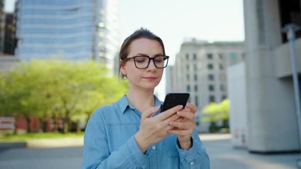 Gözlüklü beyaz kadın şehirde dolanıyor ve akıllı telefon kullanıyor. Arka planda gökdelenler var. İletişim, iş günü, yoğun yaşam konsepti. - Video, Çekim