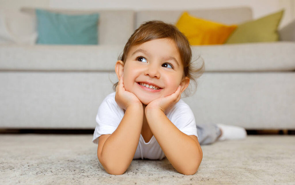 Cheerful cute little girl daydreaming on the floor at home, adorable toddler female child lying on carpet in living room, resting head on hands, smiling and looking away, copy space - Photo, Image