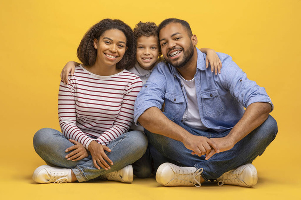 Familia afroamericana feliz de tres sentados en el suelo y abrazando, alegres padres negros amorosos posando junto con su pequeño hijo sobre el fondo amarillo del estudio, mirando a la cámara, espacio para copiar - Foto, Imagen