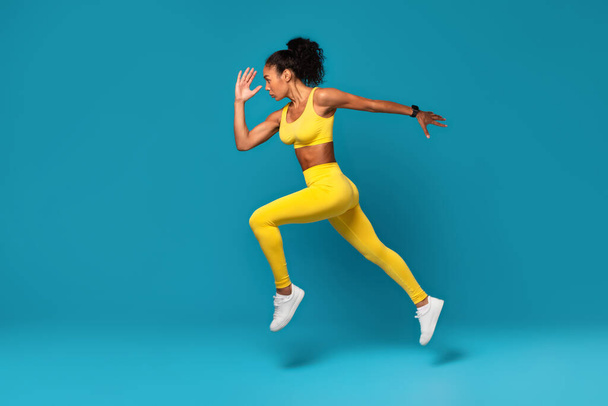 Atlética joven mujer negra en ropa deportiva amarilla saltando durante el entrenamiento dinámico en el fondo del estudio azul, epítome de la fuerza y la belleza de la aptitud. Longitud completa, vista lateral - Foto, imagen