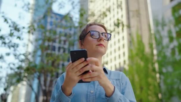 Femme caucasienne dans des lunettes est debout dans la rue dans la ville et en utilisant un smartphone. Les gratte-ciel sont à l'arrière-plan. Communication, journée de travail, concept de vie active. Mouvement lent - Séquence, vidéo