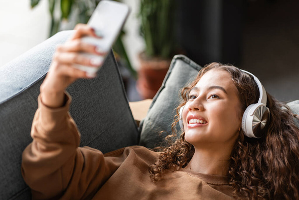 Європейська дівчина-підліток в бездротових навушниках використовує смартфон, насолоджуючись своїми вихідними з музикою та соціальними медіа, роблячи селфі, лежачи розслабленим на дивані в домашньому інтер'єрі. Гаджети, технології - Фото, зображення