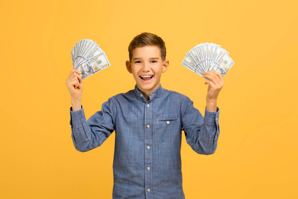 Vzrušení dospívající chlapec drží dolar v obou rukou, šťastný dospívající kluk oblečený v džínové košili stojí proti žluté pozadí studia, ilustrující bohatství, úspory, nebo finanční úspěch - Fotografie, Obrázek