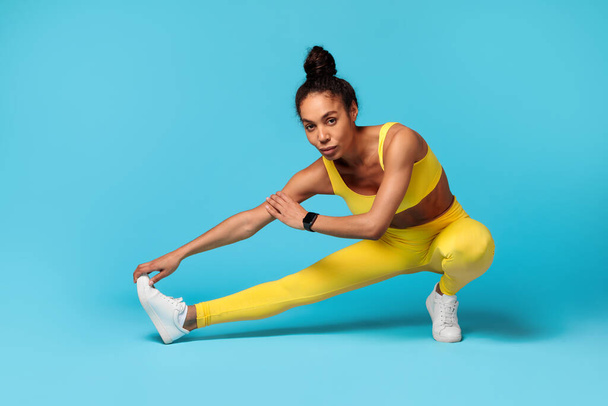 Atlética joven mujer negra haciendo ejercicio durante el entrenamiento de flexibilidad, estirando los músculos de las piernas, posando en posición de sentadilla de piernas extendidas sobre el fondo del estudio azul, mirando a la cámara. Deporte, fitness - Foto, imagen