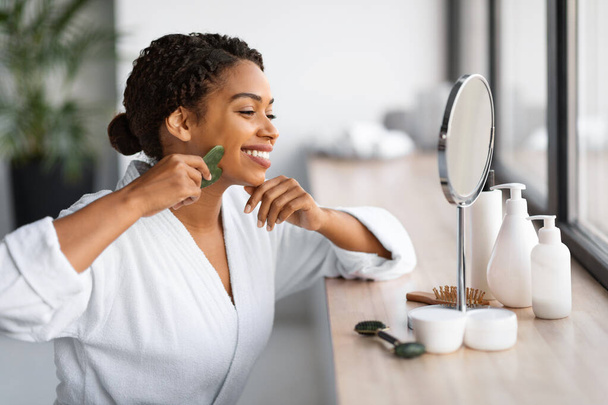 Narzędzia piękności. Uśmiechnięta młoda czarna kobieta robi masaż twarzy z Gua Sha Scraper, szczęśliwa Afroamerykanka w białym szlafroku robi zabiegi na twarz w domu, cieszy się pielęgnacją skóry, zbliżenie - Zdjęcie, obraz