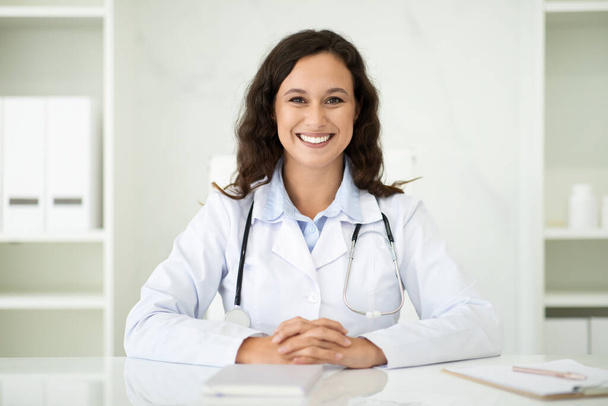Дружня тисячолітня біла жінка-лікар, сидячи за столом в інтер'єрі білого офісу в сучасній клініці, посміхається в камеру. Лікар загальної практики призначила зустріч з пацієнтом у лікарні - Фото, зображення