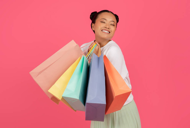 Fröhliche junge asiatische Shopaholic Frau hält helle Papiereinkaufstaschen, glückliche koreanische Frau trägt Einkäufe und schließt die Augen vor Freude, isoliert über rosa Hintergrund, Kopierraum - Foto, Bild