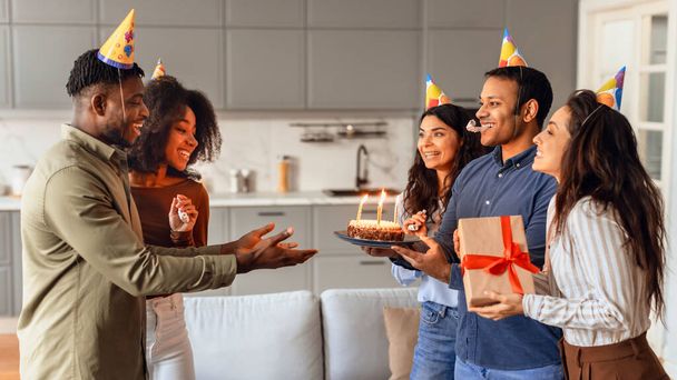 Радостная группа разнообразных друзей дарит торт на день рождения парню в праздничной шляпе, празднует день рождения и устраивает вечеринку-сюрприз в современном интерьере квартиры, преподносит подарки в гостиной. Панорама - Фото, изображение