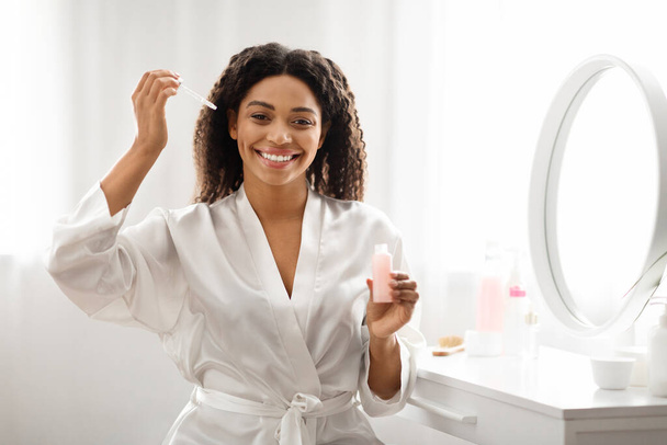 Schöne schwarze Frau trägt feuchtigkeitsspendendes Serum auf das Gesicht auf, während sie zu Hause am Schminktisch sitzt, attraktive Afroamerikanerin macht tägliche Hautpflege-Routine, lächelt in die Kamera - Foto, Bild