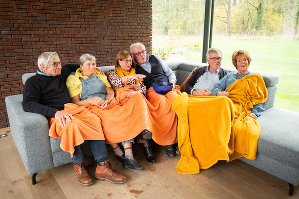 Dit beeld straalt comfort uit als een groep oudere vrienden samen ontspannen op een bank, het delen van een warme oranje deken. Hun casual kleding en de intieme setting creëren een scène van vrije tijd en - Foto, afbeelding