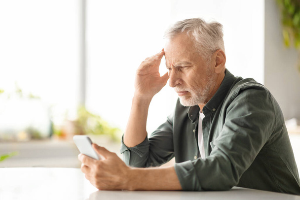 Sorunlu yaşlı bir adam evde akıllı telefona bakarken baş ağrısı çekiyor. Endişeli kıdemli beyefendi rahatsız edici haberler okuyor veya kompleks uygulamalar uyguluyor. Mutfakta masada oturuyor, uzayı kopyalıyor. - Fotoğraf, Görsel