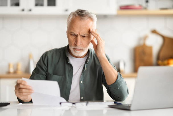 Distressed elderly man touching head, feeling overwhelmed when doing paperwork at home, senior gentleman sitting at kitchen table with laptop, reading documents and frowning, closeup - Photo, Image