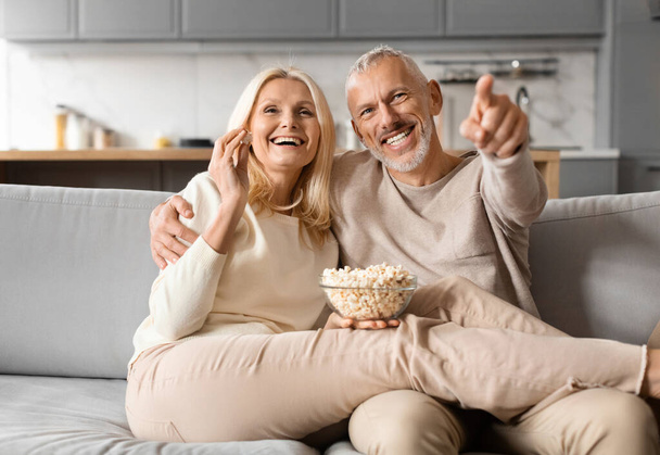 Glückliches Seniorenpaar, das zu Hause gemeinsam fernsieht. Fröhliche ältere Männer und Frauen sitzen auf der Couch, umarmen sich, essen Popcorn, das in die Kamera zeigt, genießen den Film, essen Popcorn - Foto, Bild