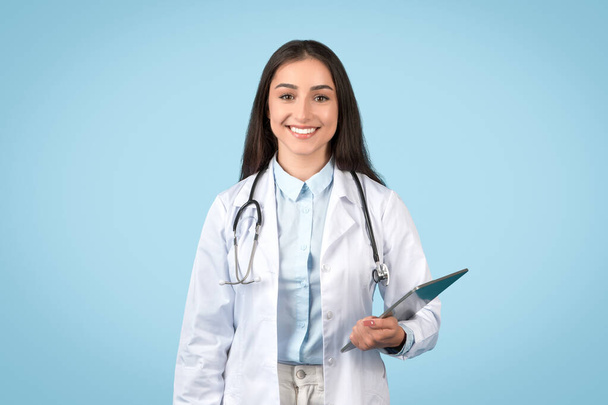 Professionelle Ärztin hält Klemmbrett für Patientennotizen bereit, mit angenehmem Lächeln und weißem Laborkittel auf ruhigem blauem Hintergrund - Foto, Bild