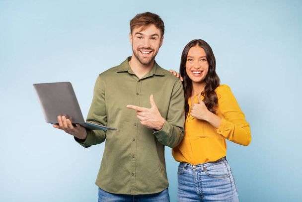 Radostný muž a žena s notebookem, muž ukazuje na počítač, zatímco žena dává palce nahoru, což naznačuje souhlas nebo úspěch na jasně modrém pozadí - Fotografie, Obrázek
