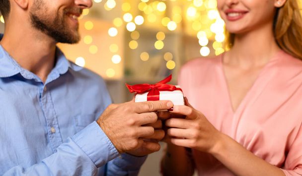 Close-up of mans hands giving white gift box with red ribbon to delighted woman, with bokeh light background adding to the romantic ambiance - Photo, Image