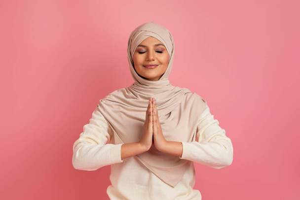 Mujer musulmana árabe alegre en hijab de pie con los ojos cerrados y las manos apretadas, encarnando la serenidad y la gratitud, posando sobre fondo de estudio rosa, simbolizando la armonía interior y el orgullo cultural - Foto, Imagen