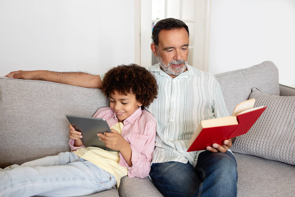 Οικογενειακός ελεύθερος χρόνος. Ισπανόφωνος Senior Man ανάγνωση του βιβλίου, ενώ ο εγγονός του χρησιμοποιώντας ψηφιακή ταμπλέτα, χαλαρώνοντας μαζί στον καναπέ στο σύγχρονο σαλόνι στο σπίτι. Διαδίκτυο και παραδοσιακό χόμπι - Φωτογραφία, εικόνα