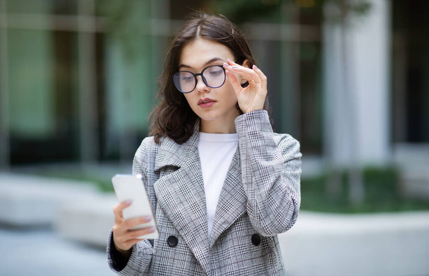 Jeune femme d'affaires européenne regardant smartphone à travers des lunettes, lire le message et à l'aide d'applications mobiles, debout sur la rue de la ville près du bâtiment moderne du centre d'affaires. Offre de communication - Photo, image
