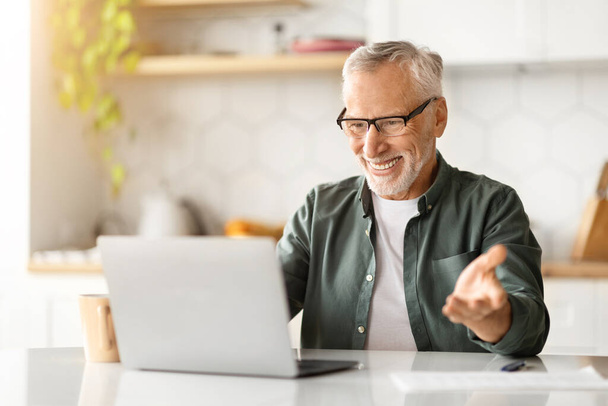 Gözlüklü neşeli yaşlı bir adam mutfaktaki dizüstü bilgisayarında video görüşmesi yapıyor, mutlu yaşlı bir beyefendi evde web kamerasında konuşuyor, arkadaşça çevrimiçi sohbetler yapıyor, uzayı kopyalıyor. - Fotoğraf, Görsel