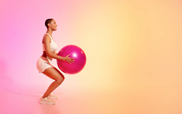 Zdolna kobieta z ogoloną głową wykonuje przysiady z różową piłką fitness, ubrana w białą odzież sportową na harmonijnym tle gradientowym. Odpowiednie, sportowe i zdrowotne - Zdjęcie, obraz