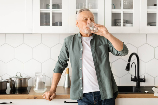 Homme âgé en bonne santé buvant un verre d'eau dans sa cuisine, homme âgé montrant l'importance de l'hydratation pour le bien-être, debout dans une posture détendue, reflétant un mode de vie sain - Photo, image