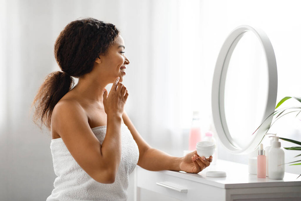 Schwarze schöne Frau, die ihr Gesicht mit Feuchtigkeitscreme eincremt, während sie zu Hause in den Spiegel schaut, afrikanisch-amerikanische Dame, die Schönheit hervorhebt, die auf Hautfeuchtigkeit und Selbstpflege zentriert ist - Foto, Bild