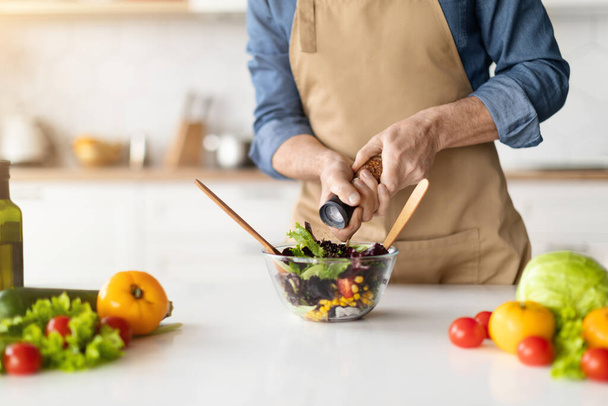 Önlüklü bir adam mutfak tezgahına serpiştirilmiş taze sebzeli renkli bir kase salata, tanınmayan bir erkek sağlıklı ev yemekleri yapıyor, yemeğe tuz ya da biber ekliyor, yakın plan. - Fotoğraf, Görsel