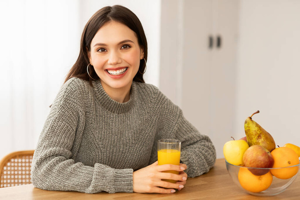 Gülümseyen esmer kadının portresi evde taze meyve suyu içiyor, mutfakta masada oturuyor, organik meyveler yiyor. Pozitif kadın sabah vitaminlerinin tadını çıkarıyor kokteyl, fotokopi alanı - Fotoğraf, Görsel
