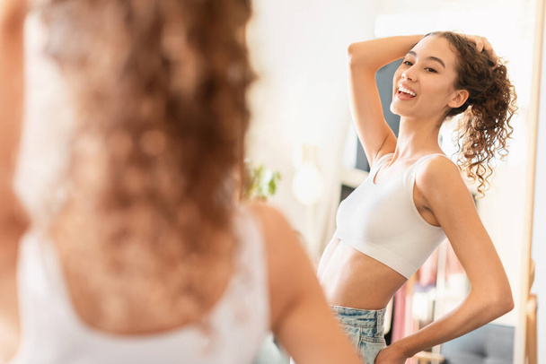 Kaukasisches Teenager-Mädchen, das vor einem Spiegel im Haus steht, mit fröhlichem Gesichtsausdruck welliges Haar berührt, lässig moderne Kleidung trägt und bewundernd fit posiert. Positives Selbstbild und Wohlbefinden - Foto, Bild