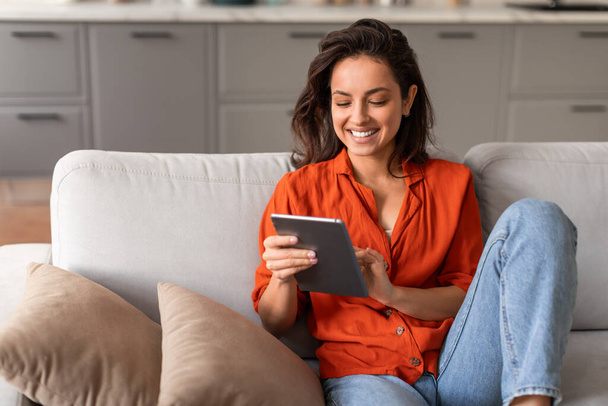 Alegre joven hembra en vibrante blusa naranja absorto en su tableta mientras cómodamente reclinado en elegante sofá gris en casa, espacio libre - Foto, imagen