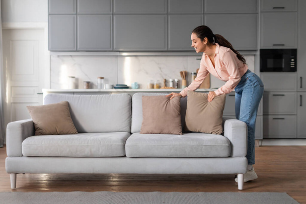 Sorgfältige Frau perfektioniert ihren Wohnraum, indem sie sorgfältig dekorative Kissen auf ihrem Sofa arrangiert, die das gepflegte und einladende Ambiente unterstreichen - Foto, Bild