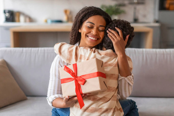 Vreugdevolle zwarte moeder met ingepakte geschenkdoos en liefdevol haar zoon omhelzend in dankbaarheid, gelukkig Afrikaans Amerikaans gezin van twee die liefdevolle momenten delen in hun gezellige woonkamer, vrije ruimte - Foto, afbeelding