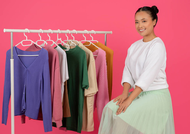Belle jeune femme asiatique assise gracieusement près du porte-vêtements et souriant à la caméra, heureuse coréenne femme contemplant des choix de mode en boutique, posant sur fond de studio rose, gros plan - Photo, image