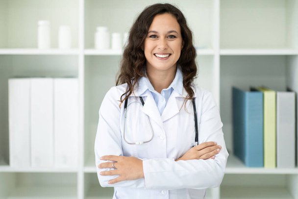 Vidám, millenniumi európai nő fehér orvosi köpenyben pózol az irodájában a modern klinikán, keresztbe tett karokkal a mellkasán, mosolyogva a kamerába. Egészségügyi koncepció - Fotó, kép