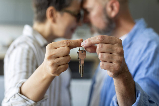 Closeup Shot Of Romantic Senior Couple Holding Home Keys, Loving Older Spouses Celebrating Moving To Their Own House, Embracing And Touching Foreheads, Cropped Image With Selective Focus - Photo, Image