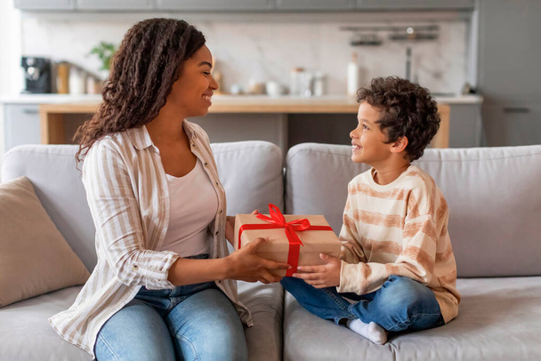 Ενθουσιασμένη μαύρη γυναίκα που λαμβάνει δώρο για την ημέρα των μητέρων από το μικρό γιο της, χαριτωμένο αφρικανικής Αμερικής αρσενικό παιδί έκπληξη μαμά με δώρο, ενώ περνούν χρόνο μαζί στο σπίτι, δωρεάν χώρο - Φωτογραφία, εικόνα