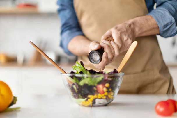 Focused elderly chef preparing nutritious vegetable salad, adding spices for flavor, unrecognizable male cooking in well-equipped kitchen, reflecting commitment to culinary passion, closeup - Photo, Image