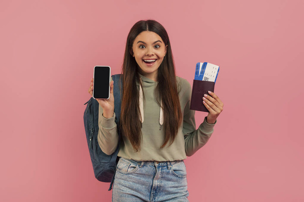 Iloinen teini-ikäinen tyttö näyttää tyhjää älypuhelinta ja pitää passia tarkastuskorteilla, iloinen teini-ikäinen nainen, jolla on reppu valmiina matkustamiseen, seisoo vaaleanpunaista taustaa vasten, johon sisältyy seikkailu - Valokuva, kuva