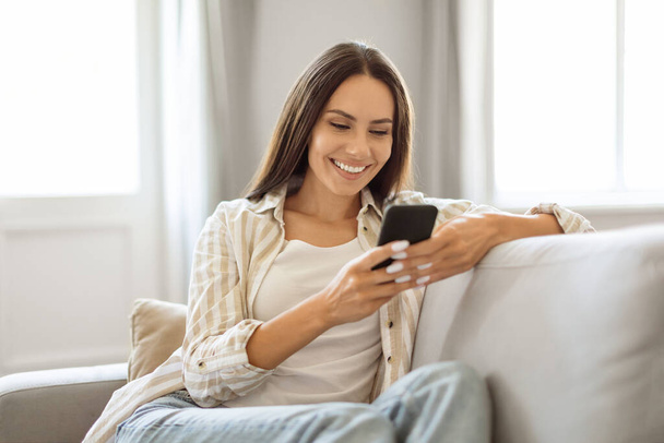 Zrelaksowana młoda kobieta uśmiechnięta i przeglądająca swój smartfon leżąc na kanapie, szczęśliwa millenium dama korzystająca z telefonu komórkowego do komunikacji online lub zakupów, odpoczywająca w salonie - Zdjęcie, obraz