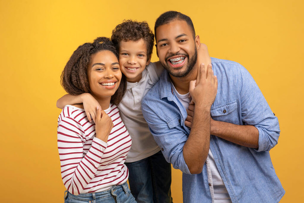 Happy African American family of three with preteen son embracing each other warmly while posing against yellow studio background, black parents and male child expressing love and togetherness - Photo, Image