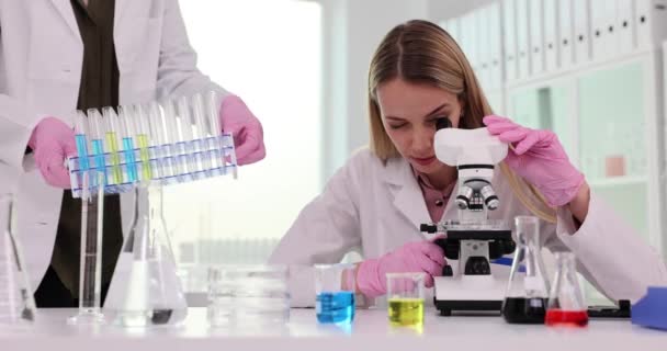 O cientista olha para o microscópio e colega coloca tubos de ensaio com líquidos coloridos. Colaboração e investigação química - Filmagem, Vídeo