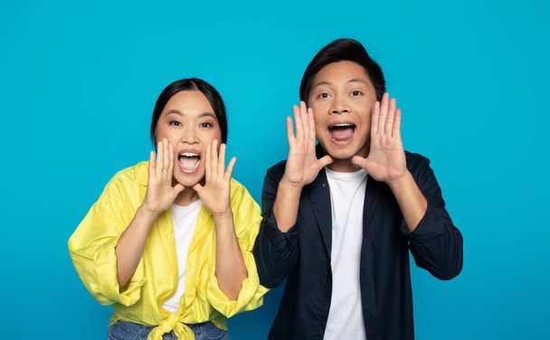 Two joyful Asian millennial friends are shouting with hands cupped around their mouths against a vibrant turquoise blue studio background, expressing excitement and fun - Photo, image
