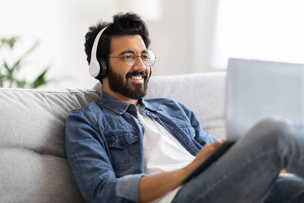Hombre independiente indio que trabaja en el ordenador portátil en línea en casa y escucha música con auriculares inalámbricos, haciendo investigación para un nuevo proyecto, sentado en el sofá con la computadora, disfrutando de un trabajo remoto, Copiar el espacio - Foto, imagen