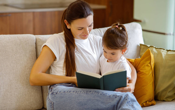 Fürsorgliche junge Mutter, die ihrer niedlichen kleinen Tochter zu Hause ein Buch vorliest, liebevolle Mutter, die sich mit dem Vorschulkind verbindet, beispielhaft für Erziehung und die Freude an mütterlicher Alphabetisierung - Foto, Bild