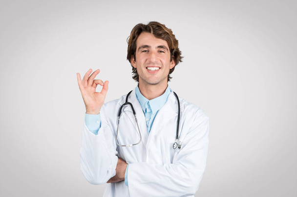 Fiducioso medico di sesso maschile con stetoscopio intorno al collo facendo gesto giusto, indicando il successo del trattamento o la soddisfazione del paziente, su sfondo grigio - Foto, immagini