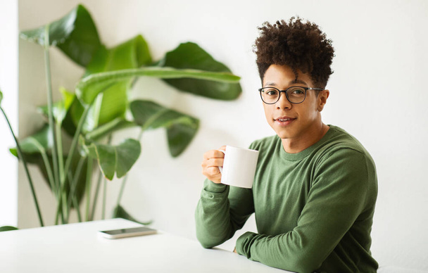 Stylish african american guy with curly hair wearing eyeglasses hipster drinking coffee at cozy cafe, sitting alone at table, smiling at camera. Leisure, coffee break concept - Photo, Image